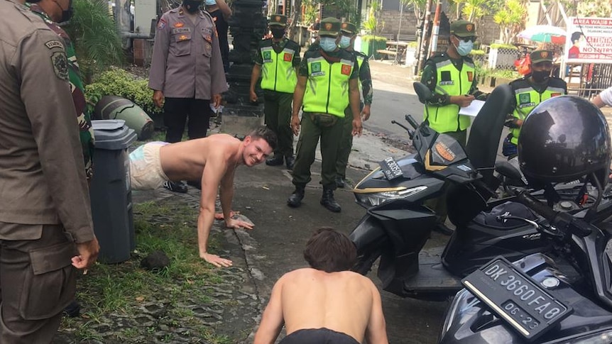 Dua pria melakukan push-up di jalan-jalan Bali dikelilingi oleh petugas polisi.