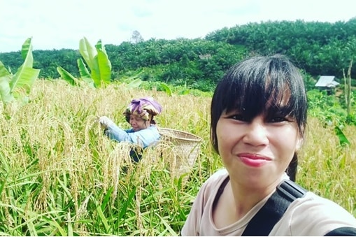 Seorang wanita mengambil foto di sawah dengan seorang petani perempuan di latar belakang.