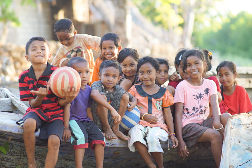 Sekelompok anak duduk memegang bola dan tersenyum di dekat pohon palem.
